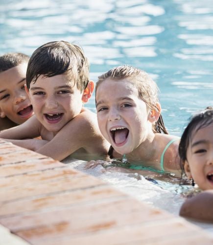Six young children laugh in a pool with their elbows resting on the pool's edge.