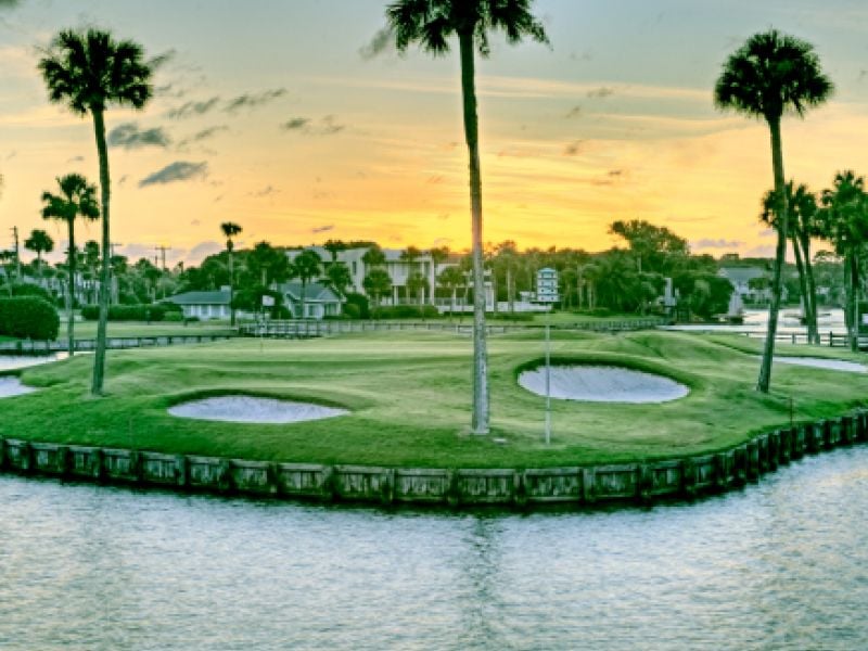 A view of the Lagoon Course at Ponte Vedra Inn & Club with lagoon in foreground, with bunkers and palm trees at sunset.