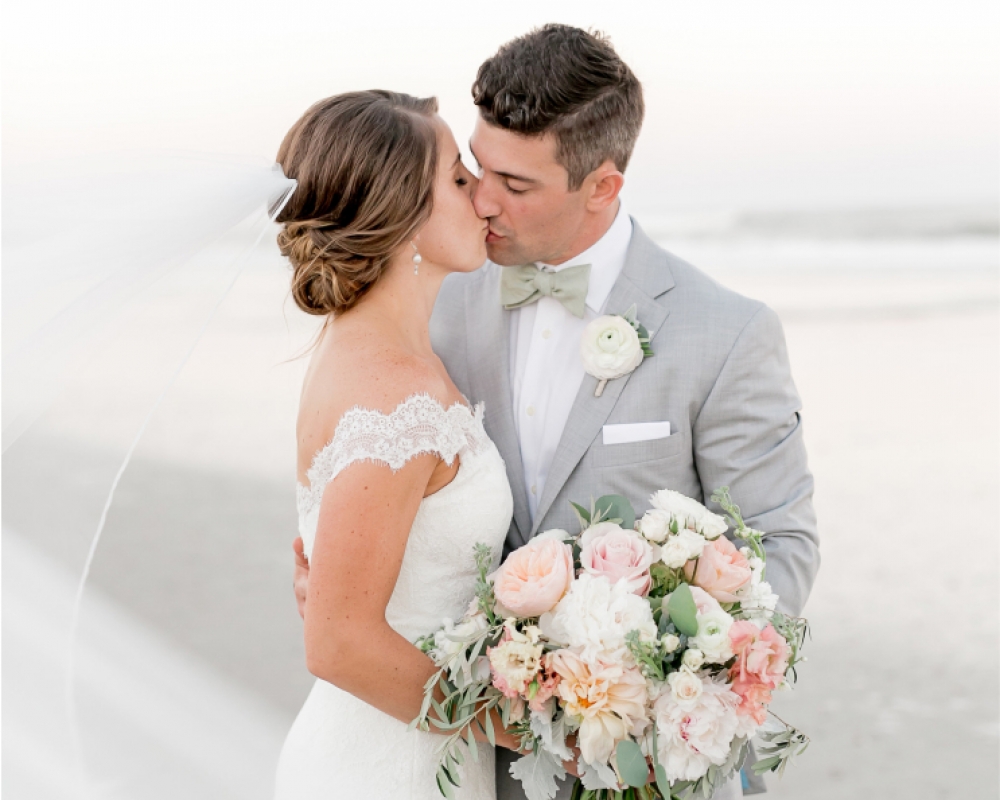 A bride and groom kiss on the beach while holding a bouquet of white and pink flowers.