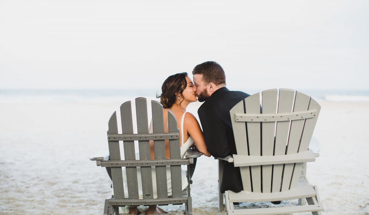 A bride and groom kiss as they sit in separate Adirondack chairs on the beach at Ponte Vedra Inn & Club. 