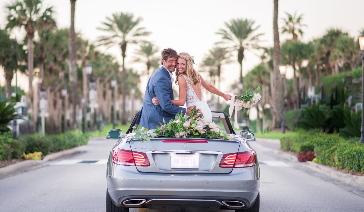 A bride and groom sit on the back of a Mercedes convertible looking behind them and smiling with a palm tree-lined street in the background.
