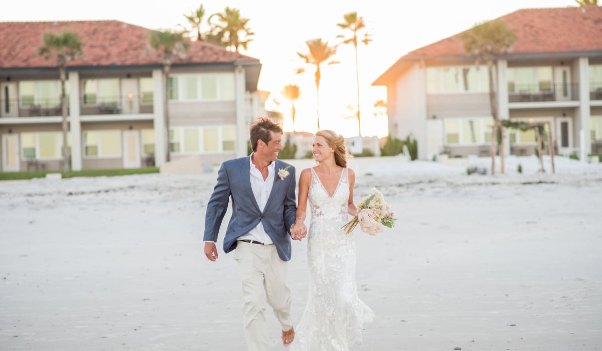 A bride and groom walk hand in hand on the beach as the sun sets behind Ponte Vedra Inn & Club.