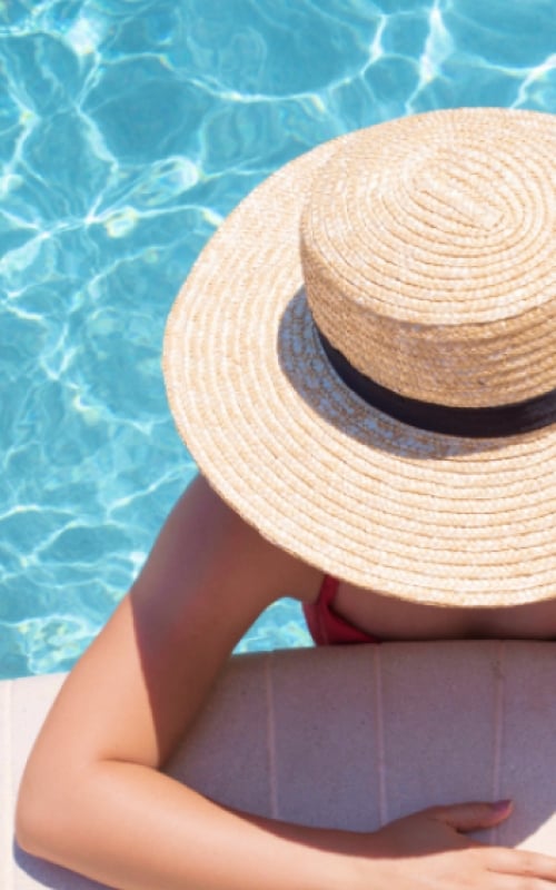 A woman stands in a pool with her arms resting on the edge and a sun hat obscuring her face.