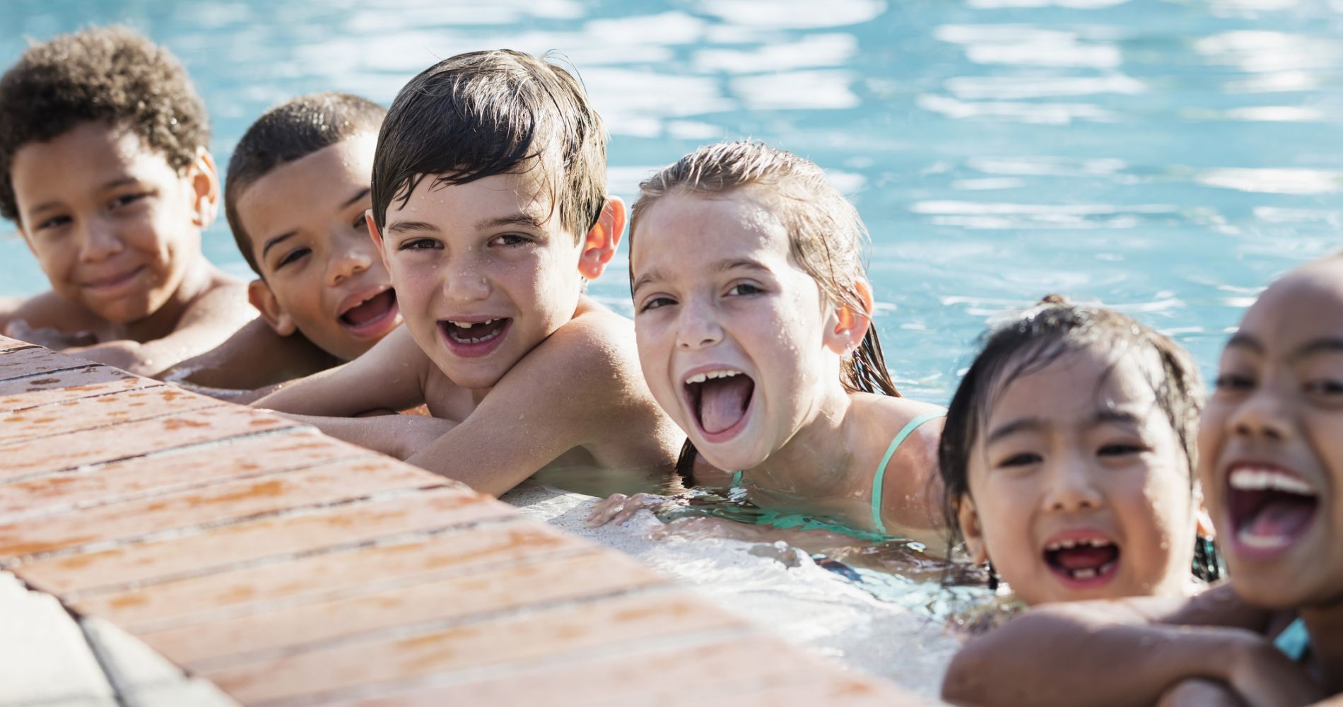 Six young children laugh in a pool with their elbows resting on the pool's edge.